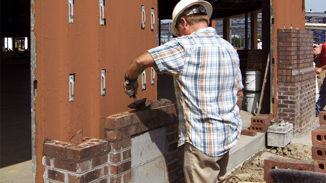 A mason lays brick over a Densglass structural wall coated with a red, fluid-applied air and water barrier. Mason contractors are discovering the benefits of installing air barriers themselves, rather than subbing it out.